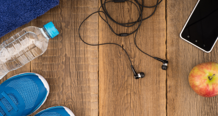 Exercise essentials: shoes, water bottle, towel, headphones, phone, and apple on a wood floor.